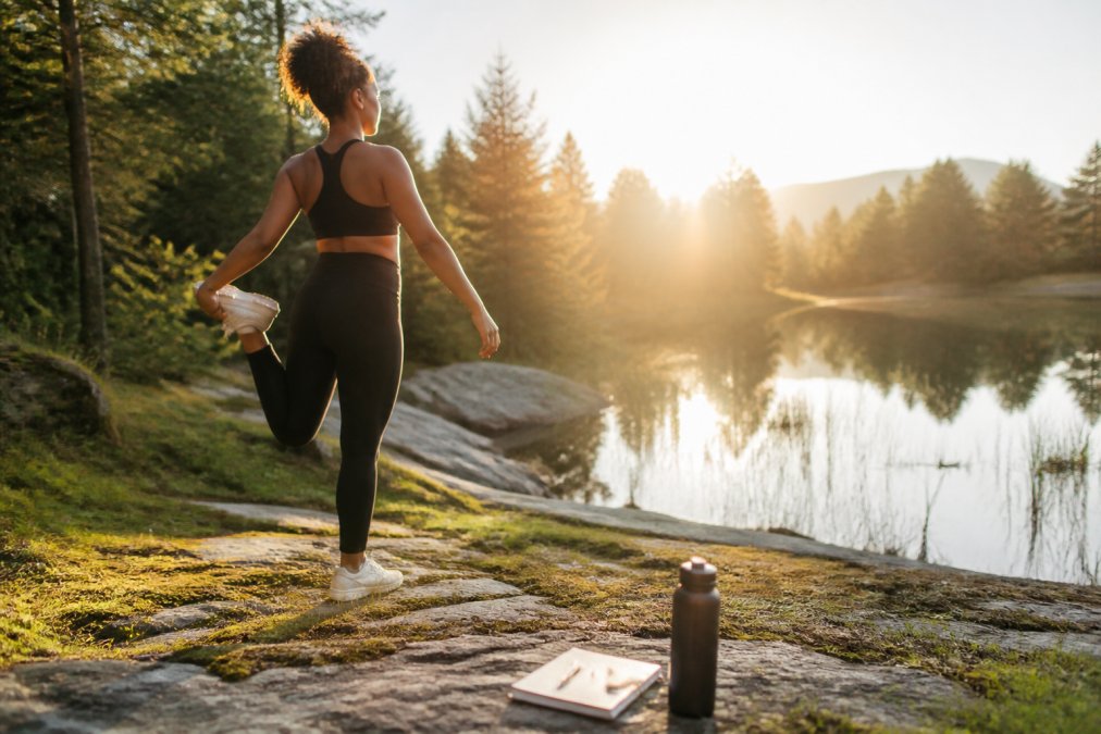 A person outdoors during morning light, stretching or walking in nature, appearing calm and energized with a water bottle and journal nearby
