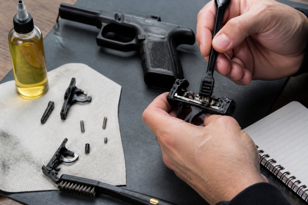 Close-up of a hand using a small brush to clean the trigger assembly of a striker-fired pistol on a workbench, with cleaning solvent and a maintenance log notebook visible nearby