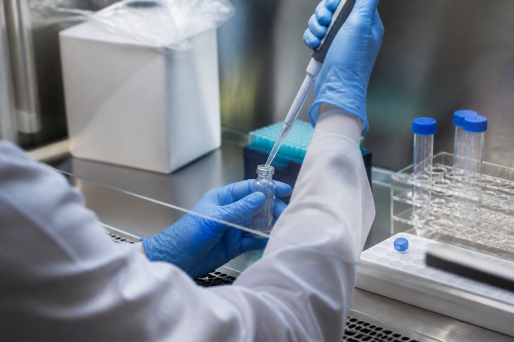 Researcher's gloved hands carefully reconstituting lyophilized peptide powder with pipette into buffer solution in small vial under controlled laboratory hood with precise lighting