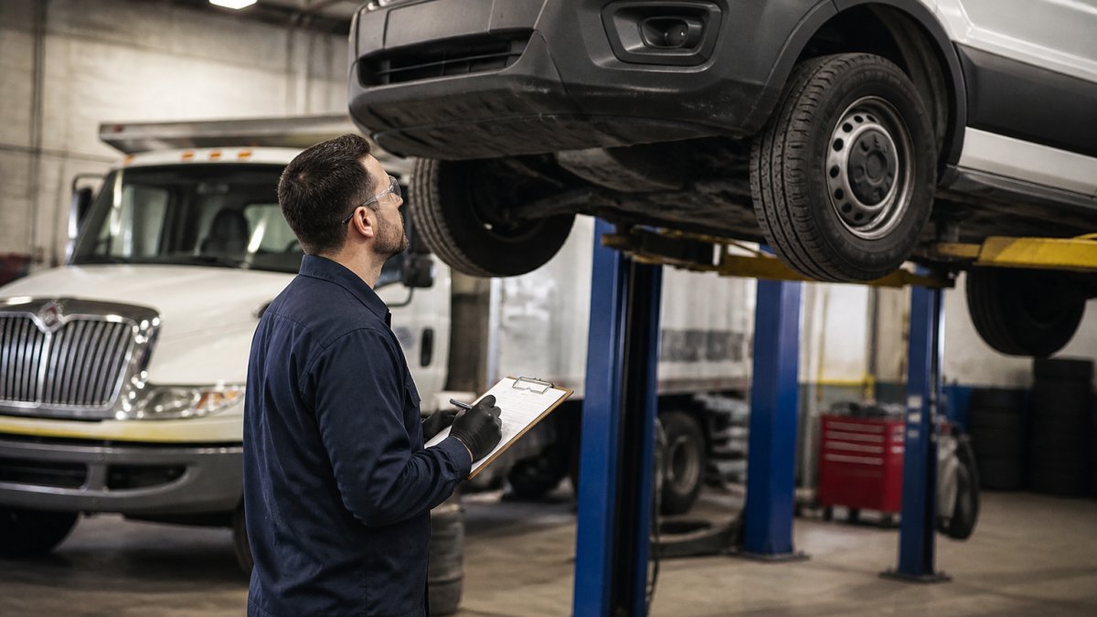 An ASE-certified technician in a clean shop bay inspecting a commercial van raised on a lift, clipboard checklist in hand, another fleet truck parked behind under bright overhead lighting