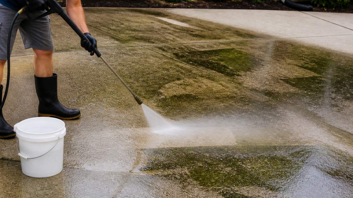 Close-up of a homeowner using a low-pressure washer to clean green algae and dark mold spots from a concrete driveway, with a bucket of eco-friendly cleaner nearby