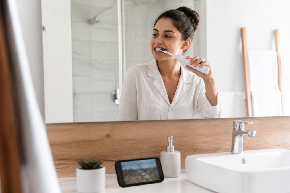 A person in a bright, modern bathroom smiling while using an electric toothbrush, with a phone playing a video on the counter nearby