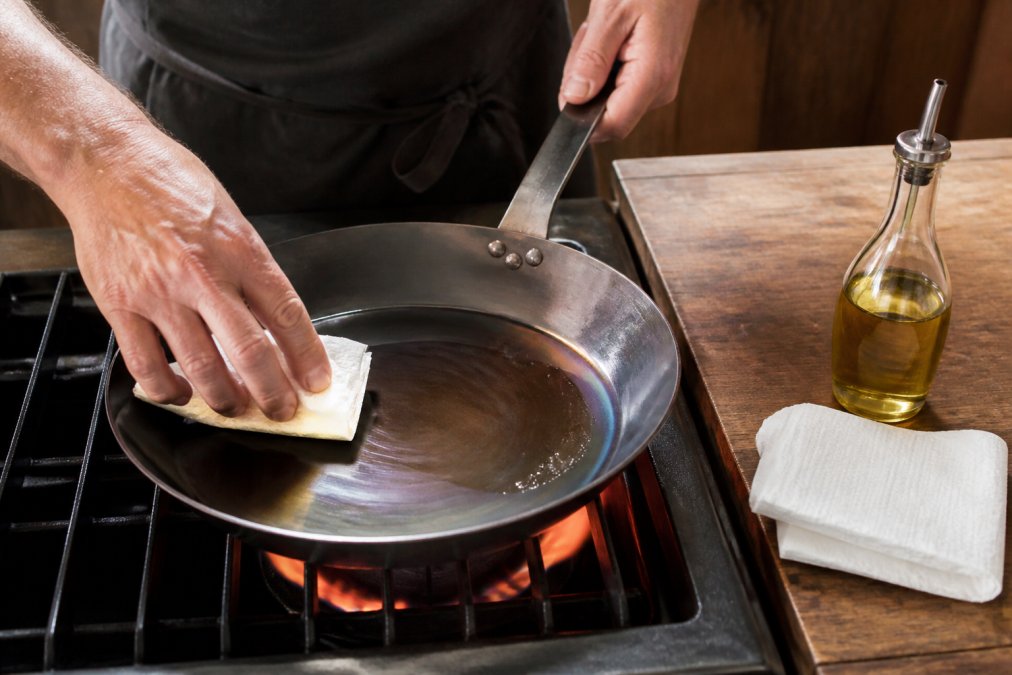 Close-up of hands applying a thin layer of neutral oil to a carbon steel skillet on a glowing stovetop burner, a folded paper towel and small glass oil bottle resting on a wooden counter beside the pan, warm kitchen lighting