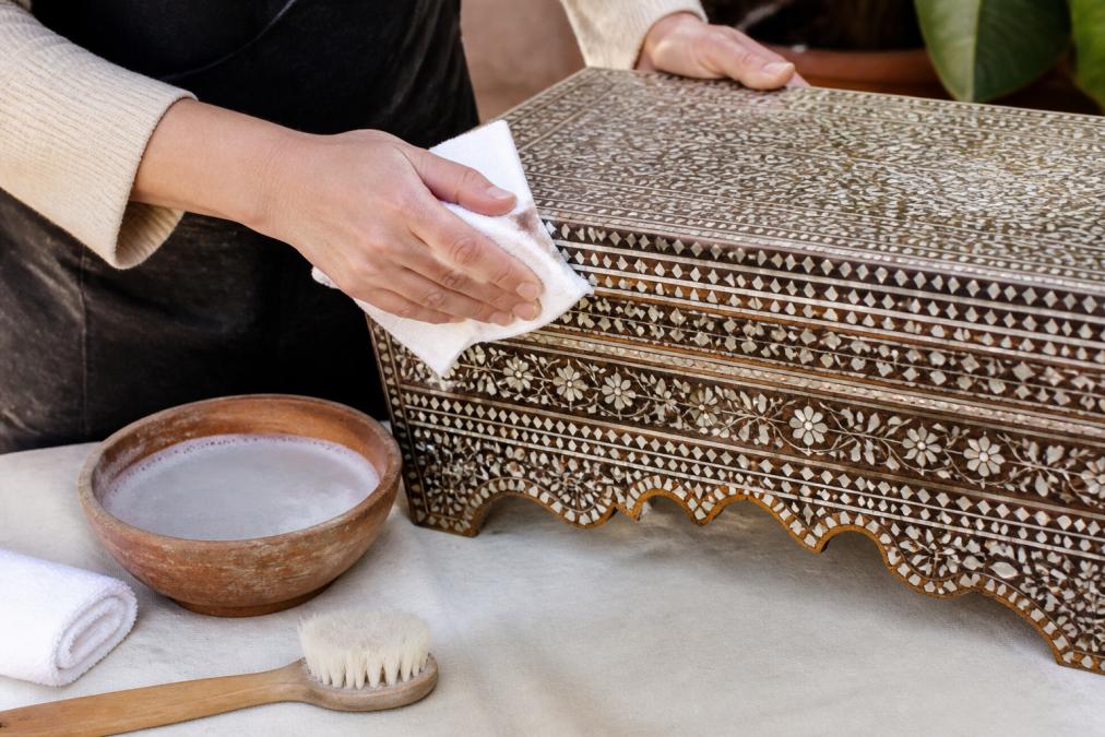Close-up of hands using a soft microfiber cloth to gently clean an ornate Moroccan mother-of-pearl inlaid wooden furniture piece, with a small bowl of mild solution nearby on a wooden surface