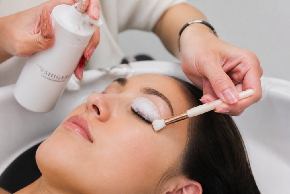 Close-up of hands gently cleansing eyelash extensions with a foaming cleanser and soft brush over a sink, showing proper technique with warm water and careful, deliberate movements