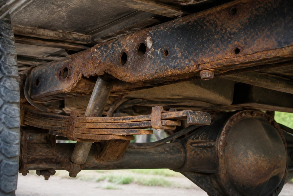 Close-up of truck undercarriage showing rust and corrosion on frame rails and suspension components, natural outdoor lighting