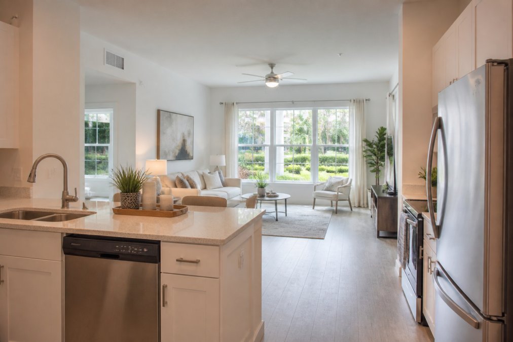 Bright, modern one-bedroom apartment interior with stainless steel appliances, quartz countertops, natural light streaming through large windows overlooking landscaped grounds