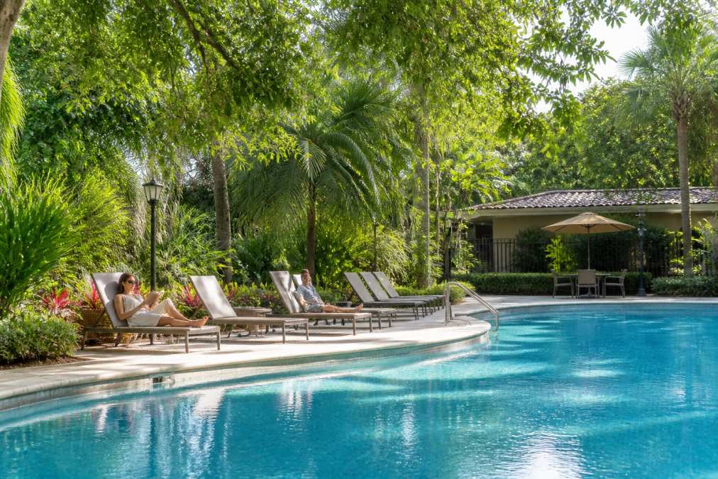 Residents relaxing by resort-style pool surrounded by lush landscaping and lounge chairs in garden-style community setting, natural sunlight filtering through mature trees