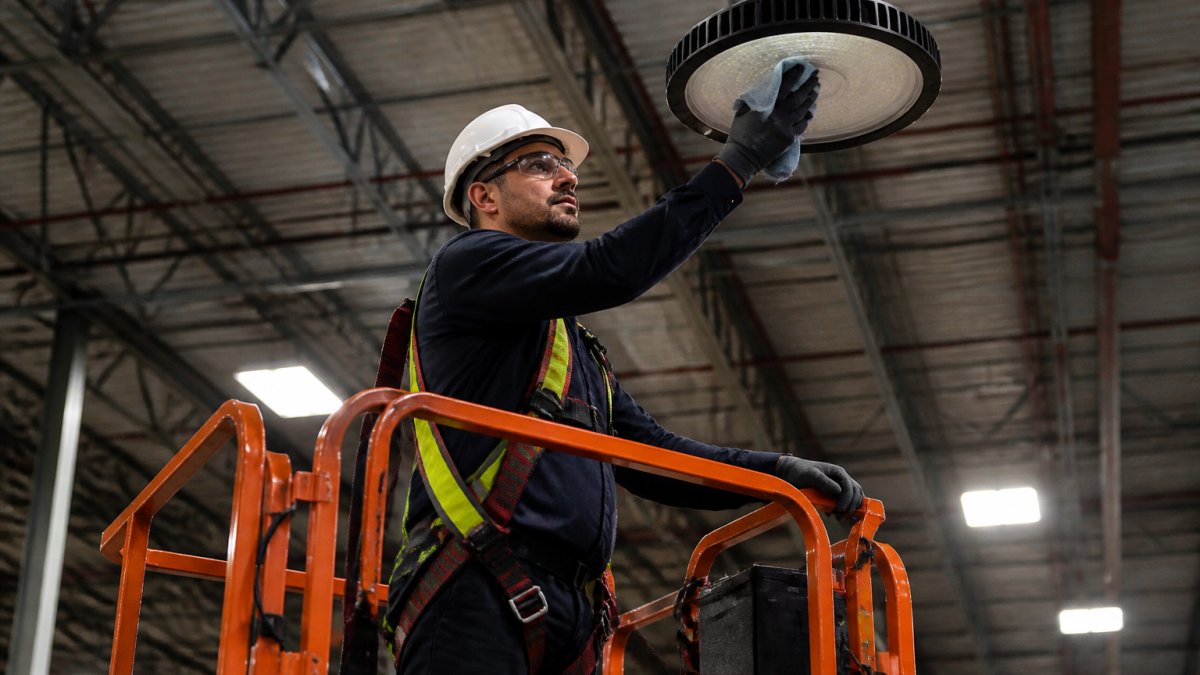 A facility maintenance technician on a scissor lift carefully wiping the lens of a high bay LED fixture with a soft cloth in a warehouse, wearing safety glasses and a hard hat