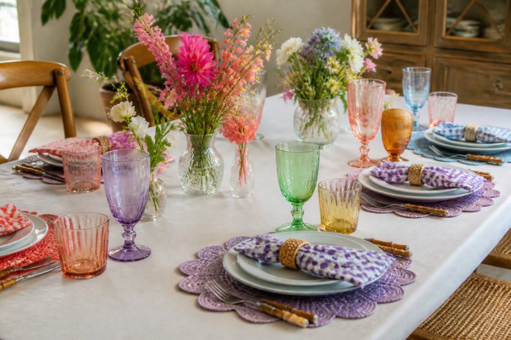Elegant table setting for entertaining featuring colorful glassware in various shapes, coordinated with patterned placemats and cloth napkins, showing how different pieces work together with dinnerware and fresh flowers