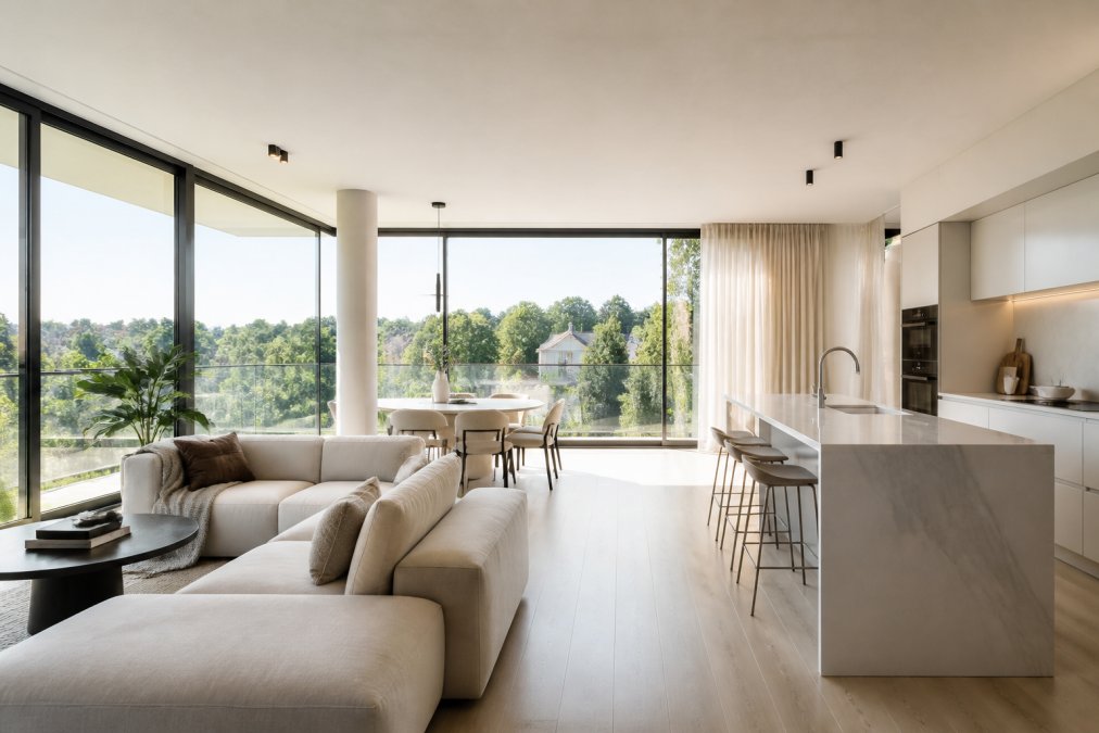 Intérieur lumineux d'un appartement moderne avec cuisine ouverte, grandes fenêtres panoramiques, mobilier contemporain minimaliste et vue sur un quartier résidentiel verdoyant d'Uccle