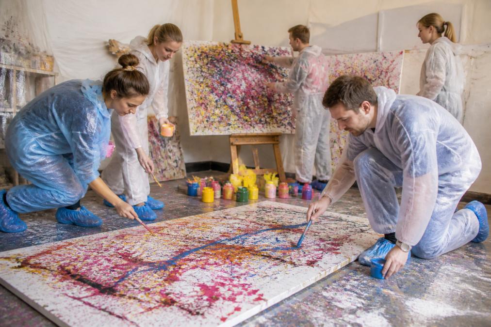 Gruppe von Teilnehmern beim Action Painting Workshop, die gemeinsam farbenfrohe Leinwände mit Pinseln und Farben gestalten, mit Farbspritzern und kreativer Energie in einer inspirierenden Atelieratmosphäre