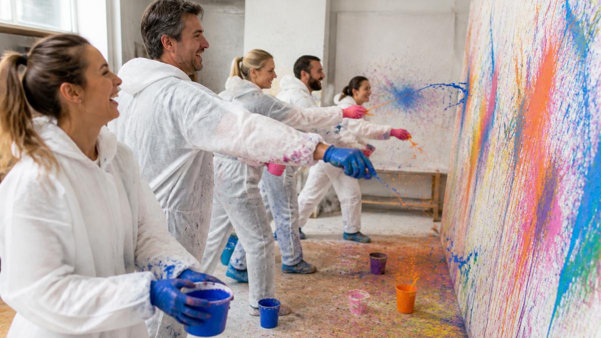 Eine Gruppe Erwachsener in Schutzkleidung wirft Farbe auf eine große Leinwand in einem hellen Atelier, lachend und mit Farbspritzern auf dem Boden