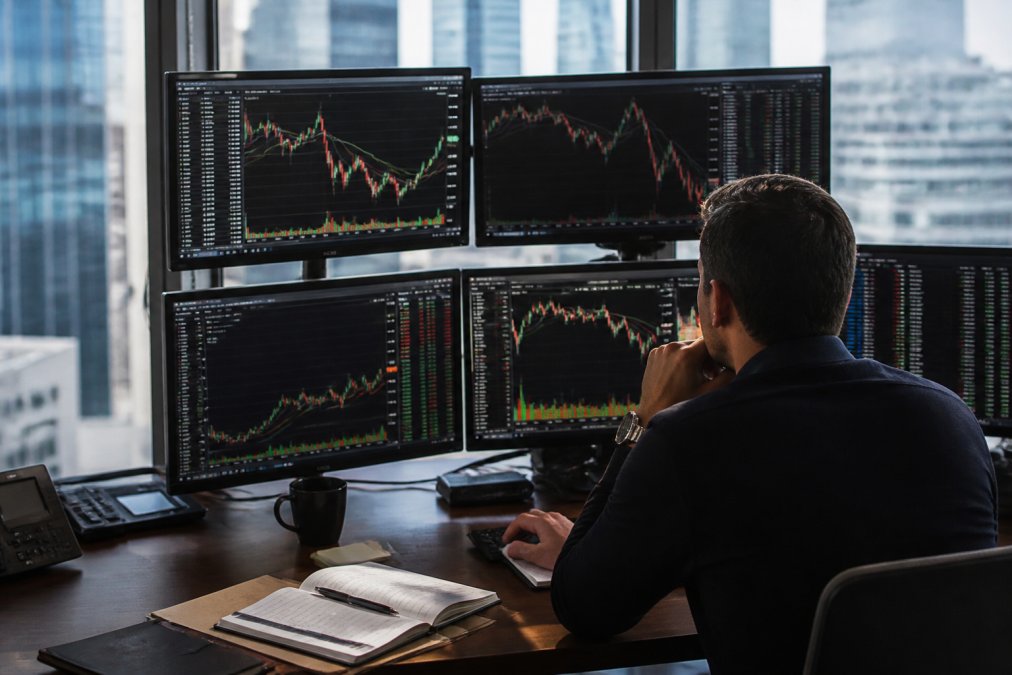 Professional trader seated at desk with multiple monitors displaying live market data, candlestick charts, and real-time trading platforms, with coffee and trading journal nearby