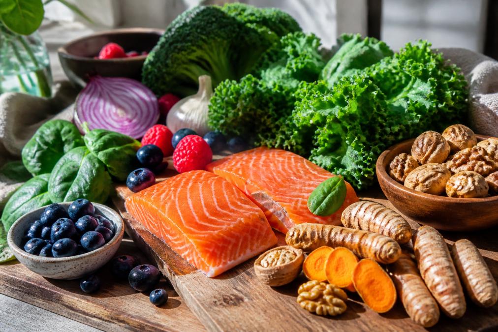 Colorful spread of fresh vegetables including leafy greens, bright berries, fatty fish fillets, walnuts, and turmeric root arranged on a wooden cutting board with natural morning sunlight