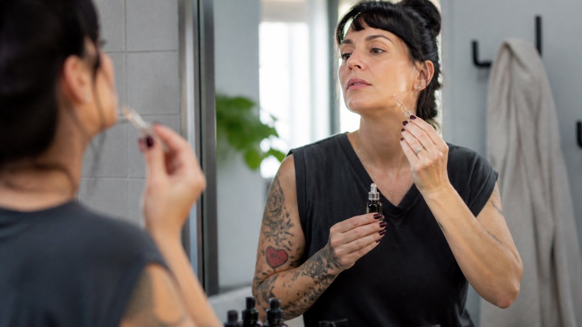 Woman in her 40s applying serum to her chin in morning light, looking at herself in a bathroom mirror with confidence