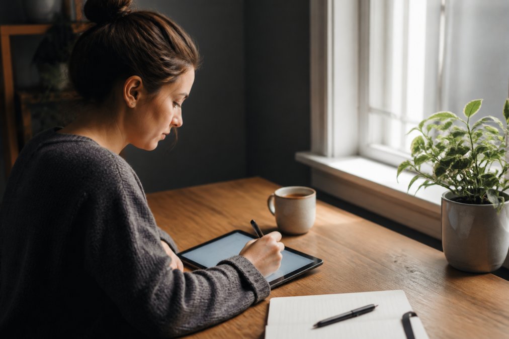 Person sitting at a desk with a cup of coffee, writing in a digital tracker on a tablet with a focused, calm expression, natural window lighting streaming across the workspace