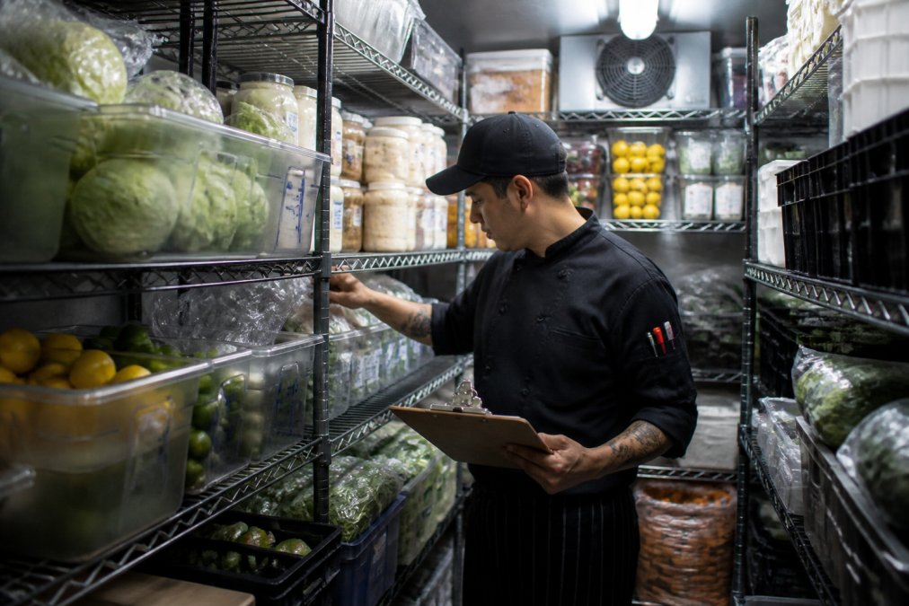 Restaurant kitchen staff checking inventory on shelves and in storage, scanning items and recording counts on a clipboard while examining perishable goods under bright overhead lighting