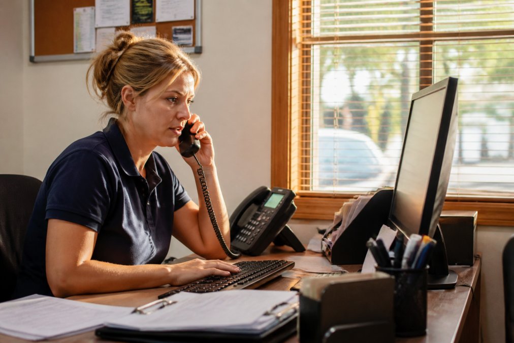 A busy receptionist in a small plumbing office answering a landline while glancing at a computer monitor that displays a queue of missed calls, warm afternoon light through a window