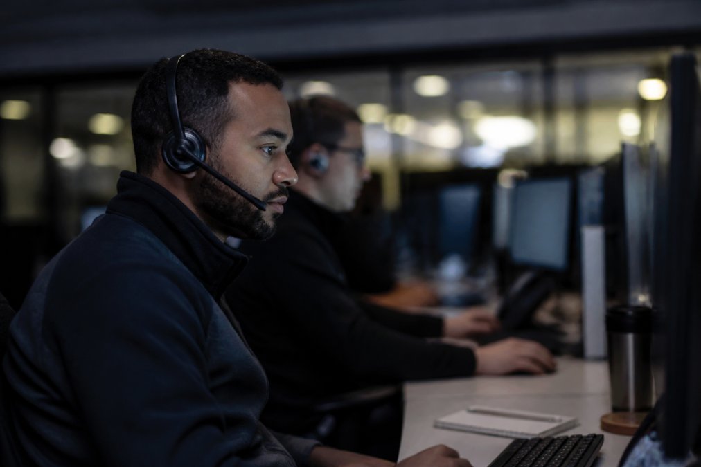 A security analyst wearing a headset monitoring multiple screens displaying network traffic and threat alerts in a dimly lit security operations center