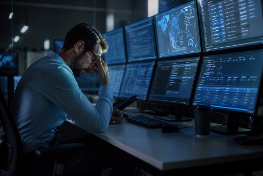 Stressed IT security professional monitoring multiple screens with security alerts and dashboards in a dimly lit control room late at night, focused on detecting threats