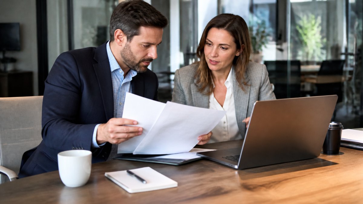 Two business professionals reviewing contract documents and a laptop at a conference table in a bright office, with a notepad and coffee nearby