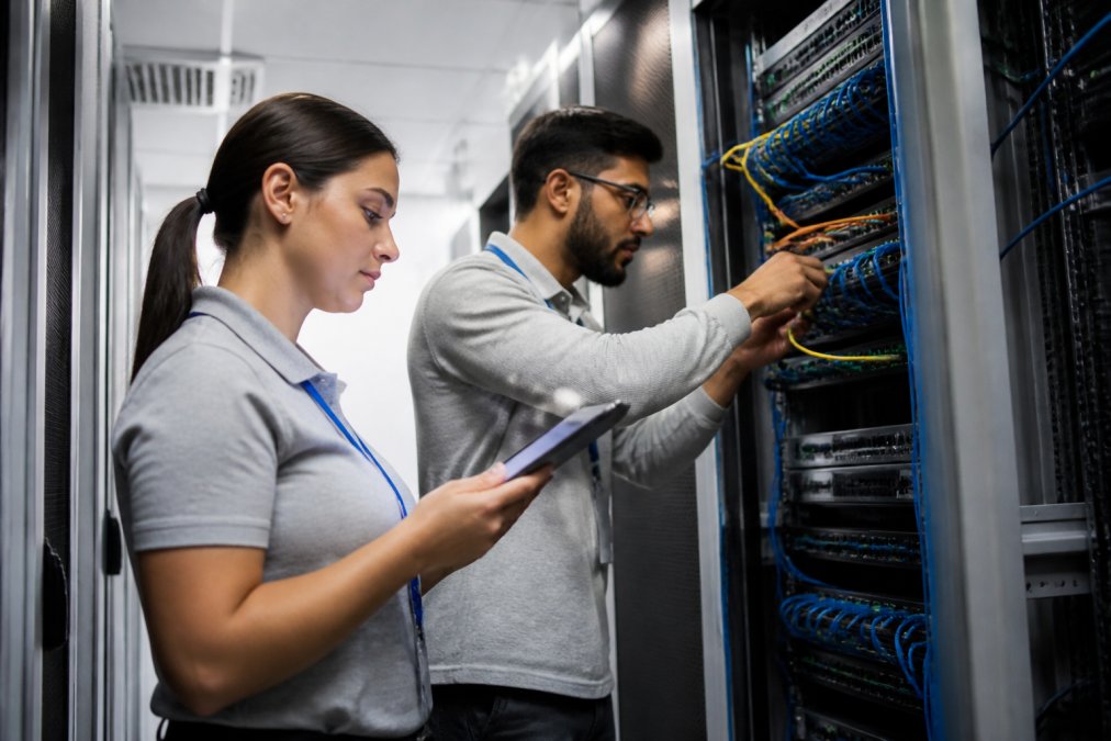 IT technician in a server room connecting ethernet cables while a colleague reviews a checklist on a tablet, suggesting an active network migration in progress with rack-mounted servers and cable management visible under cool blue-white LED lighting