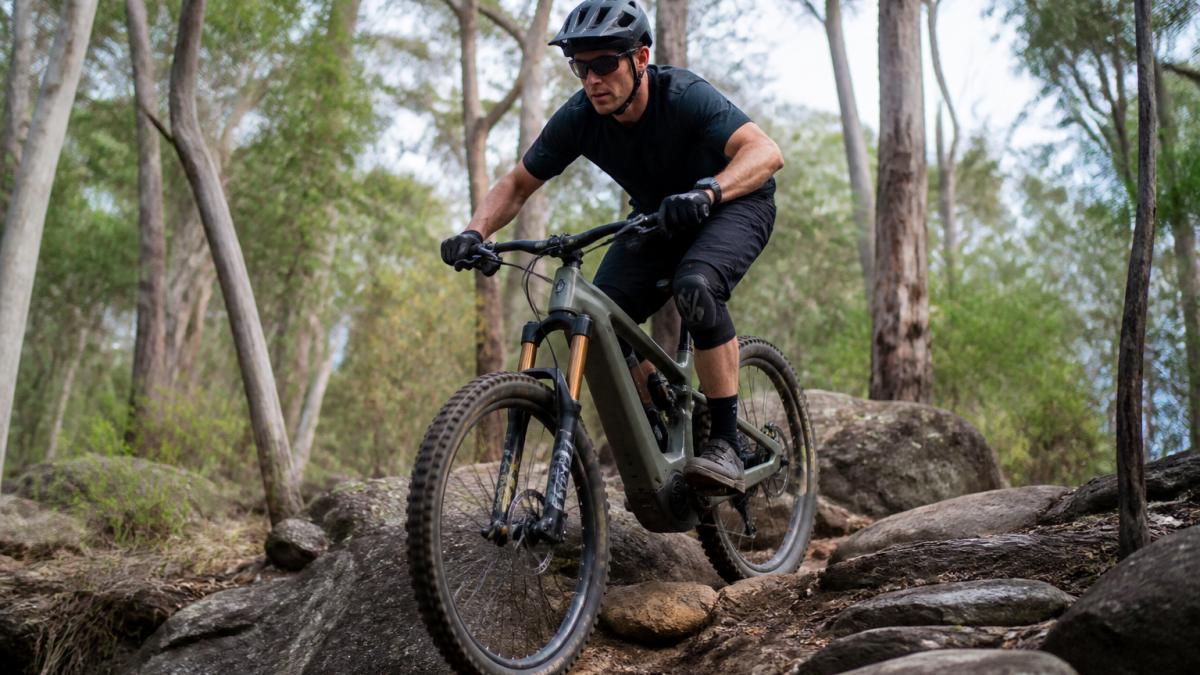A rider standing on the pedals of a full-suspension electric mountain bike on a rocky trail, elbows out and weight centred, jarrah forest in the background