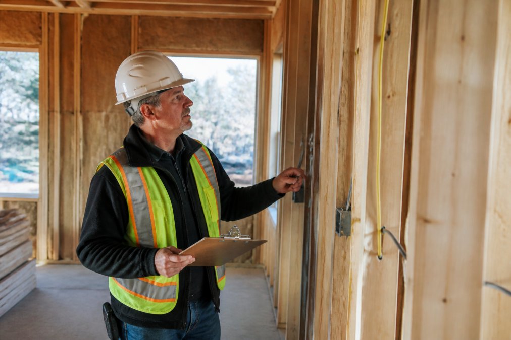 A site supervisor in a hard hat and work vest holding a clipboard, conducting a quality control inspection inside a partially finished residential room, examining wall framing and rough electrical wiring in warm afternoon light filtering through unfinished window openings