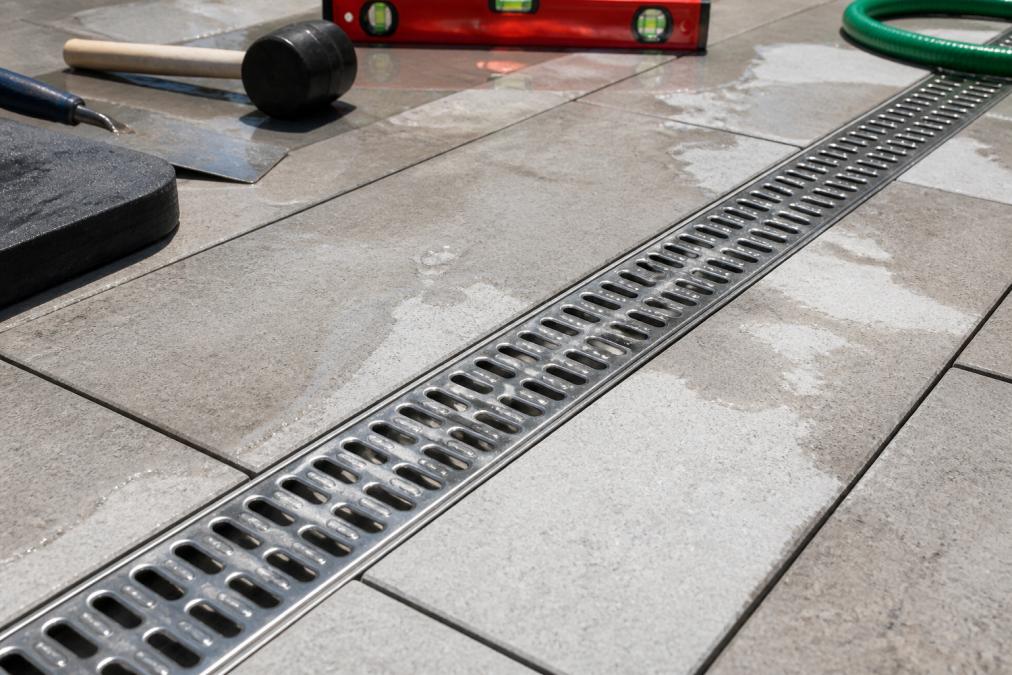 Close-up of a stainless steel channel drain grate installed flush with light gray concrete pavers on a residential outdoor patio, with a thin stream of water flowing through the grate slots, photographed from above in bright afternoon sunlight