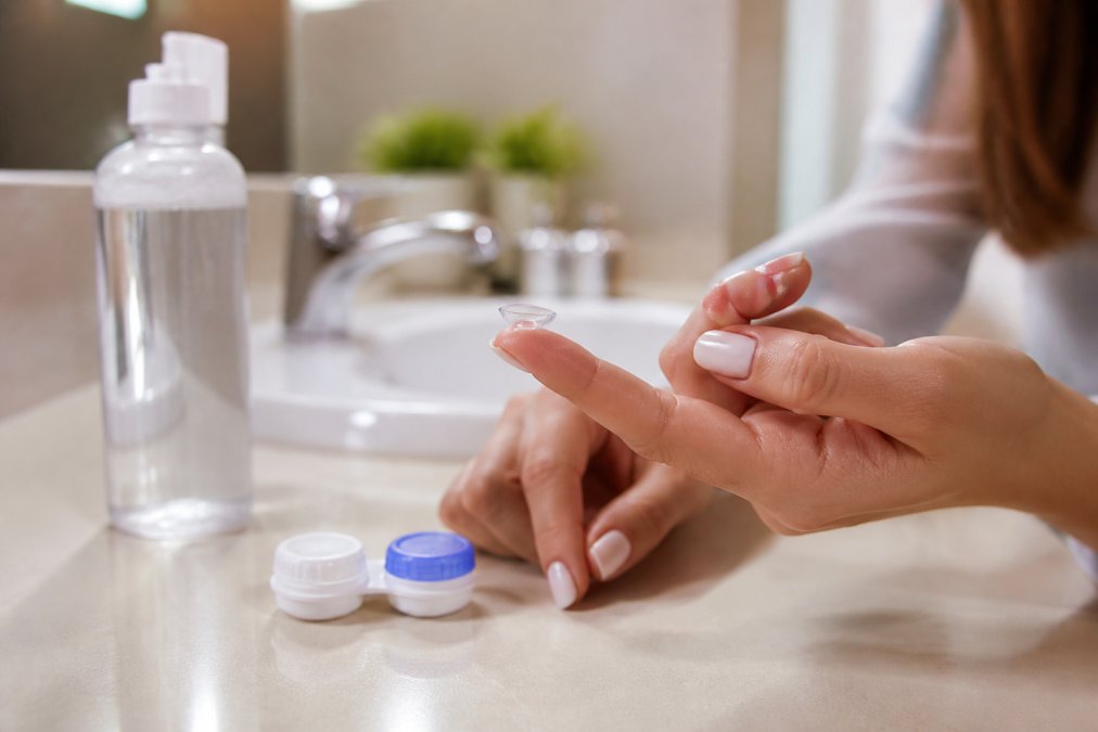 Person holding a contact lens on their fingertip while managing dry eyes with contact lenses in a bathroom.