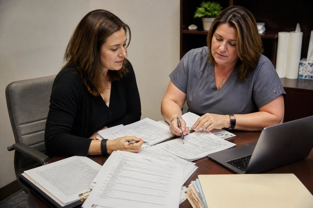 Practice manager and clinical staff reviewing patient intake forms and workflow documents at a desk with multiple papers, a laptop, and intake checklists visible under office lighting