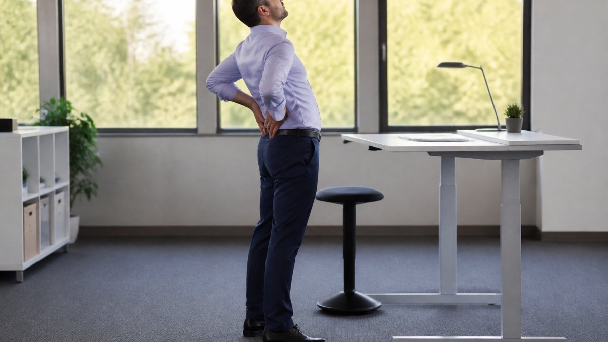 A person working at a standing desk in a bright office, taking a break to stretch their lower back with hands on hips, a small stool nearby