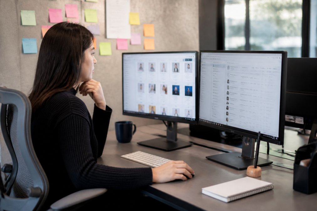 A marketing coordinator at a desk with two monitors showing campaign dashboards, sticky notes on the wall behind, coffee cup nearby, bright modern office setting