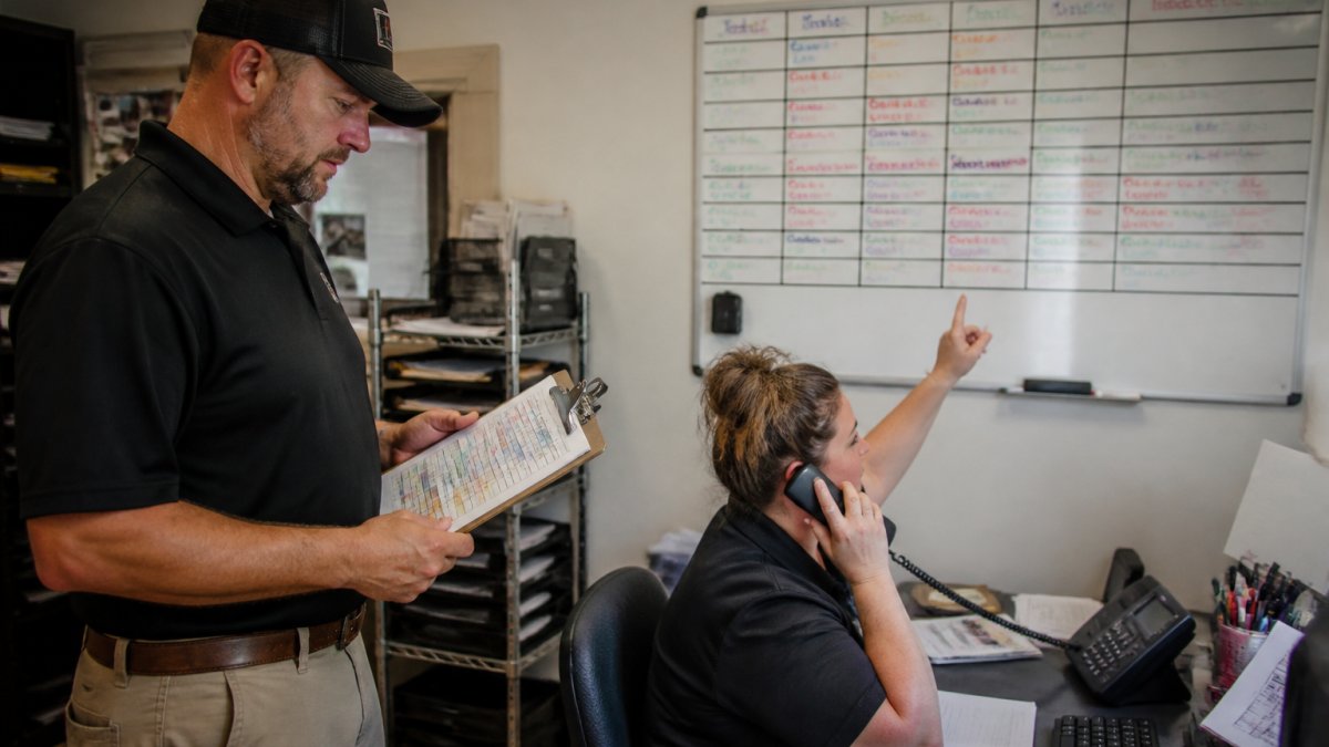 A home service branch manager standing in a dispatch office, reviewing a printed schedule on a clipboard while a dispatcher at a desk takes a call and points at a wall-mounted whiteboard of job assignments