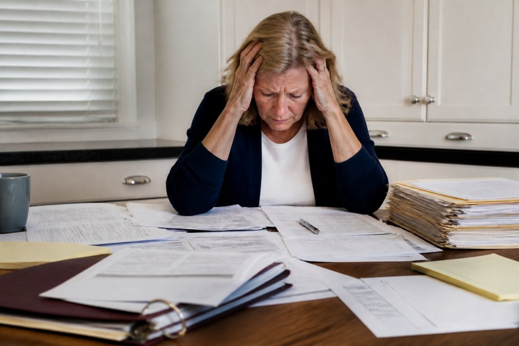 Person looking stressed while holding their head at a desk surrounded by scattered probate documents, files, and financial statements, showing overwhelm during estate administration under harsh fluorescent lighting