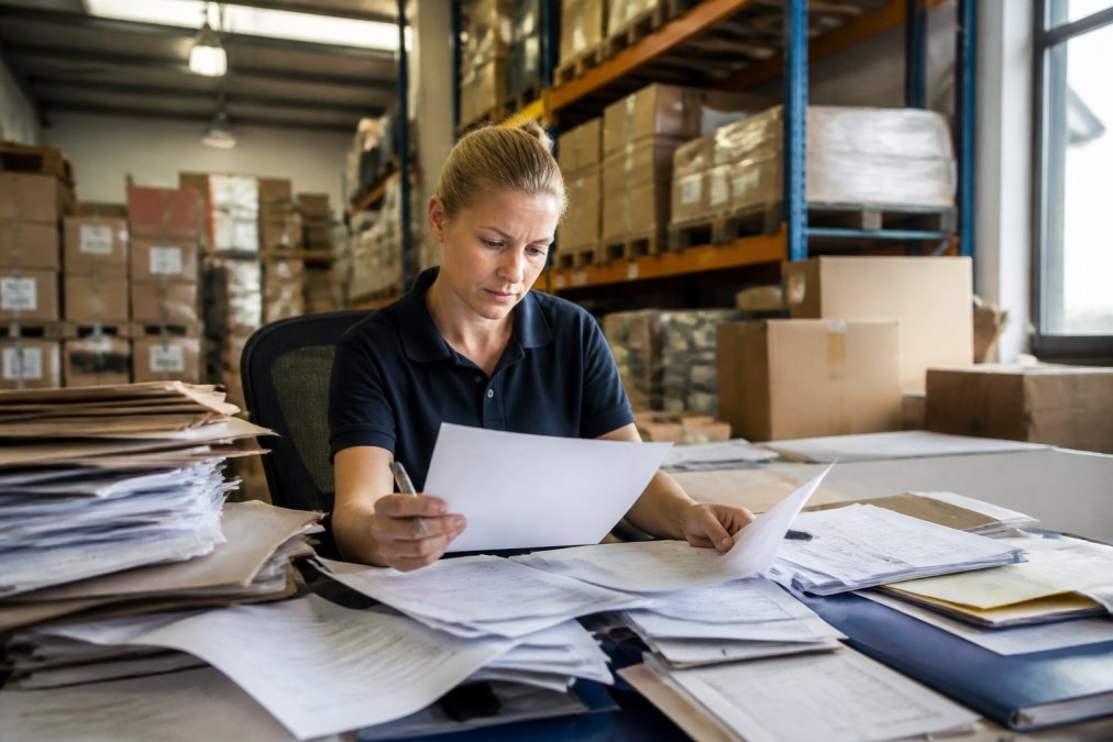 A logistics coordinator sorting through paper documents and shipping files at a cluttered desk in a busy warehouse office, natural daylight from a window