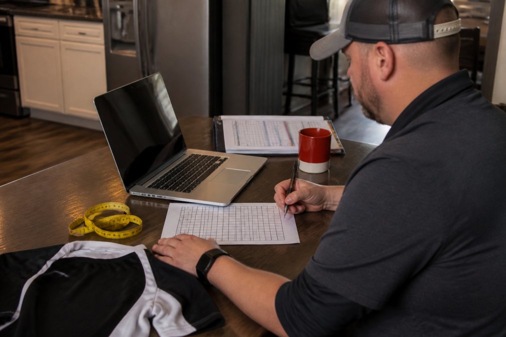 A youth sports coach at a table with a laptop open to a spreadsheet, a printed roster, a tape measure, and a sample jersey laid out beside a coffee mug