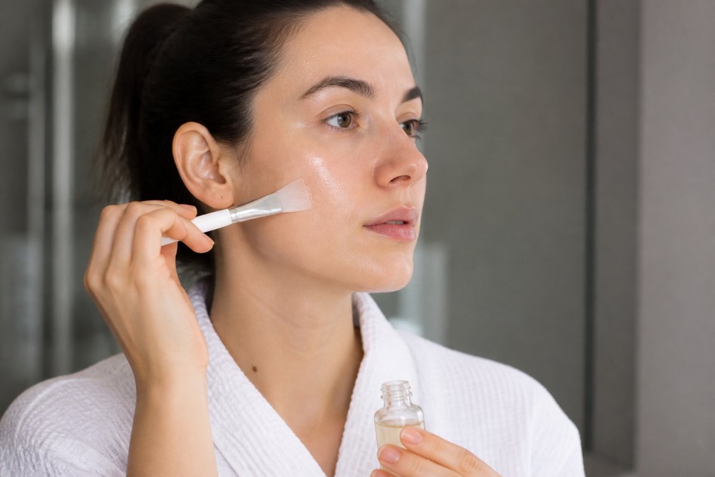 Close-up of hands applying clear mandelic acid solution to clean facial skin with a brush applicator in well-lit bathroom setting