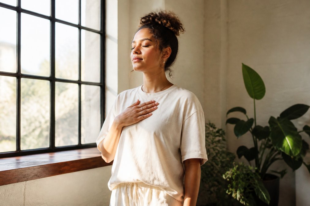 Spiritual woman wearing a manifestation tee standing in natural sunlight near a window, hand placed over heart, eyes closed in peaceful intention-setting moment