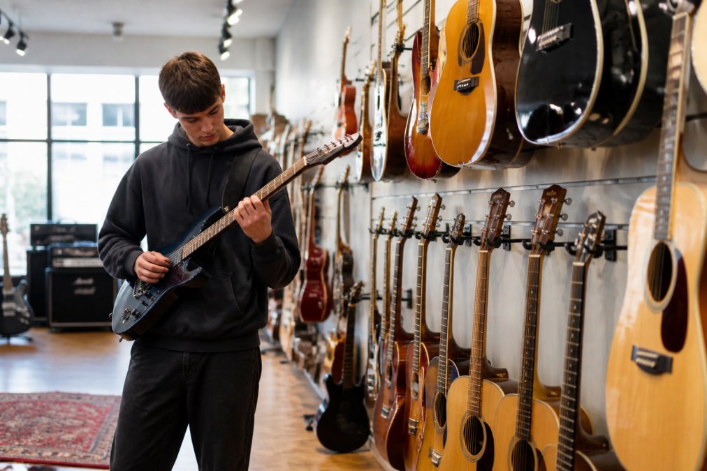 A beginner musician browsing electric and acoustic guitars displayed on wall stands in a bright, modern music shop with natural lighting from large windows, examining the fretboard of a guitar