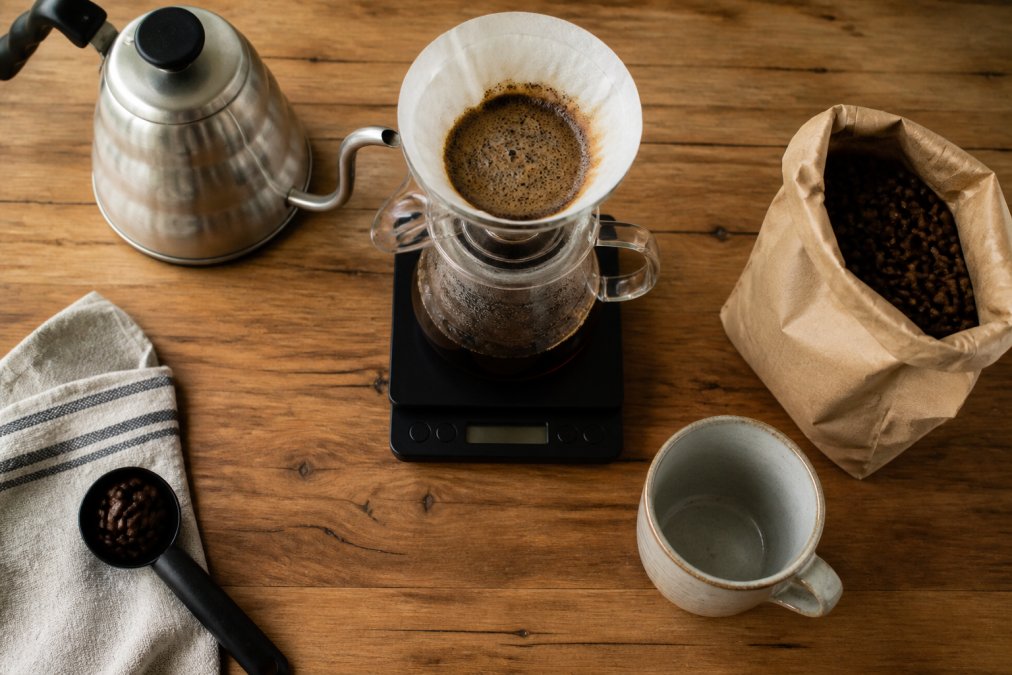 Overhead shot of a pour-over setup on a wooden counter: gooseneck kettle, glass carafe, digital scale, and a bag of whole bean coffee beside a ceramic mug