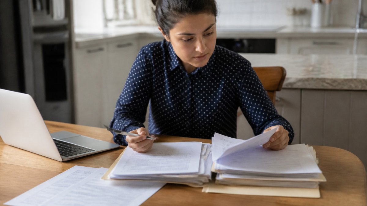 A person at a kitchen table organizing a folder of financial paperwork, including tax returns and bank statements, with a laptop open beside them and a pen in hand
