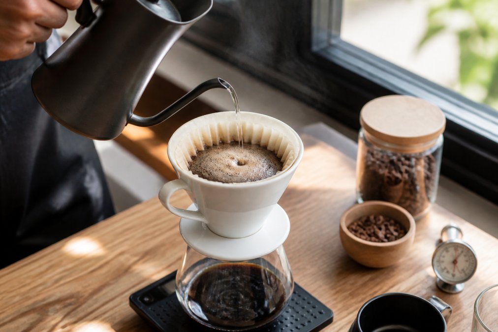 Close-up of hands pouring hot water in slow circular motions over medium-fine coffee grounds in a ceramic pour-over dripper, with specialty coffee beans and a thermometer visible on a wooden brewing station with natural window light