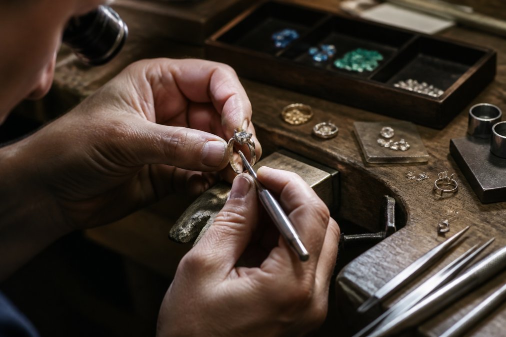 A master jeweller's hands carefully setting a diamond into a bespoke engagement ring at a precision workbench, with gemstones, metal samples, and specialised tools arranged in natural studio lighting