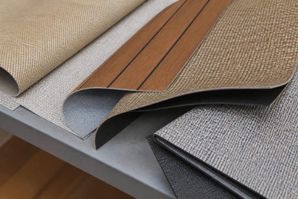 Close-up of woven vinyl flooring samples in marine-grade colors including sea grass and teak-look patterns with different backing types visible on a professional display table