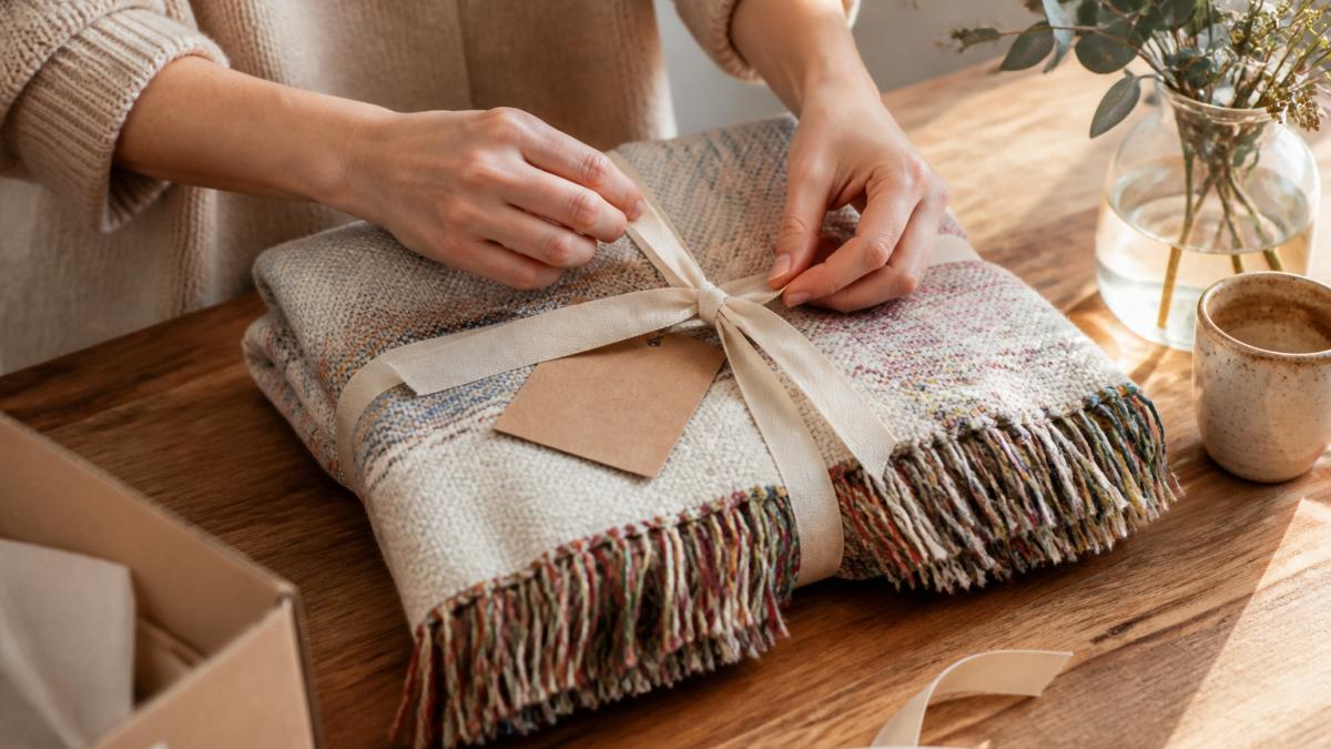 A person's hands tying a ribbon around a woven cotton blanket with colorful fringe, a small handwritten note card resting on top, warm natural light on a wooden table