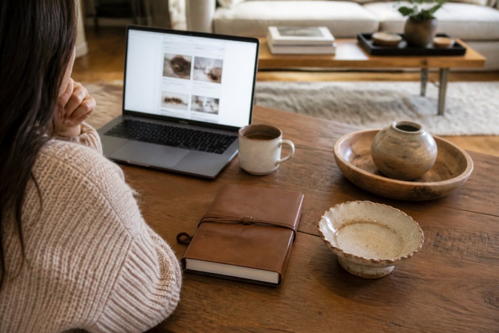 A thoughtful gift-giver comparing two personalized items on a table, a monogrammed leather journal and a handcrafted home accent, with a laptop open to a gift website in a warm, softly lit living room