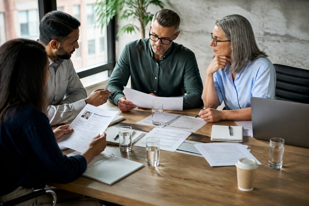 Business team reviewing candidate profiles and marketing executive resumes at a desk with laptops, notepads, and coffee, natural office lighting