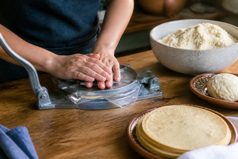 Hands pressing masa harina dough in a tortilla press on a wooden kitchen bench with cooked tortillas nearby.